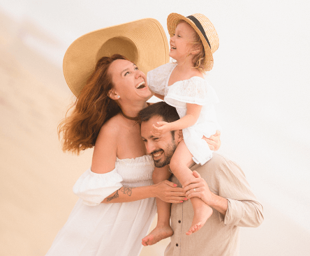 Rodina s ženou, mužem a dítětem se smějí společně na pláži. Dítě sedí muži na ramenou.