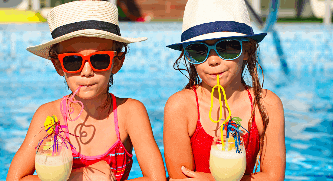 Dvě děti v bazénu popíjející shake brčky. Mají na sobě červené plavky, brýle a slaměné kobouky.