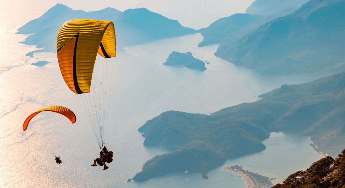 Dva lidé provozující paragliding a v pozadí jsou vidět moře a hroy.